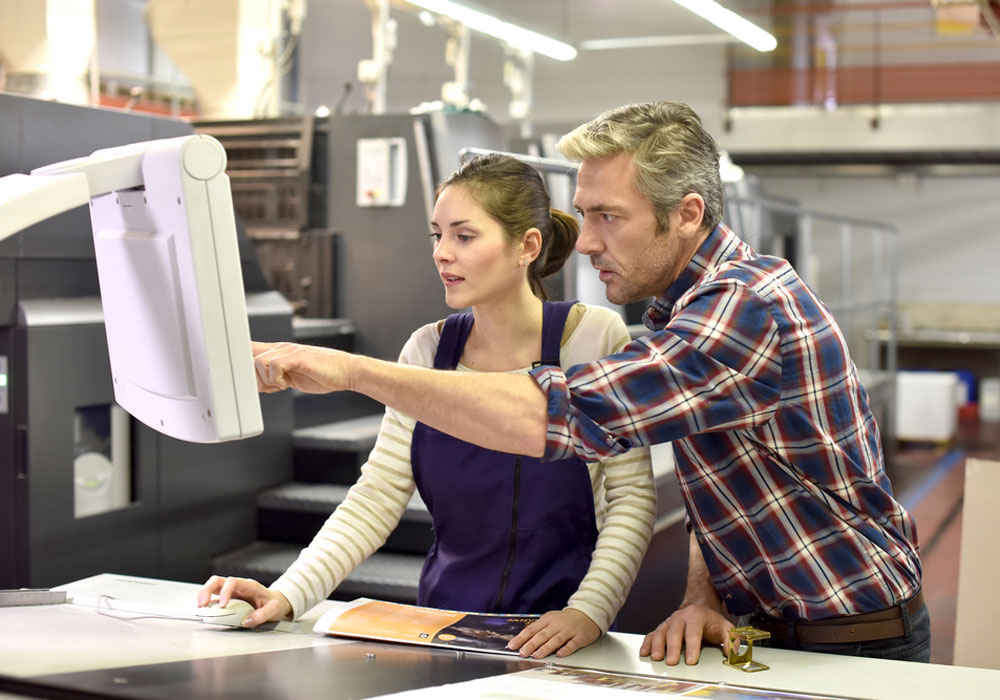 Middle aged White manager shows young white female apprentice how to work a machine in a print shop