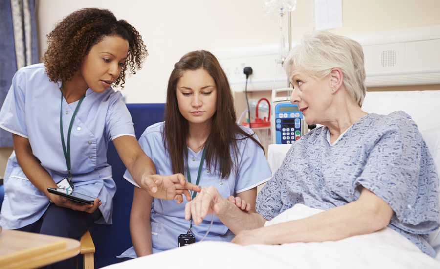 A female nurse works with a female nurse trainee to help an older white female patient
