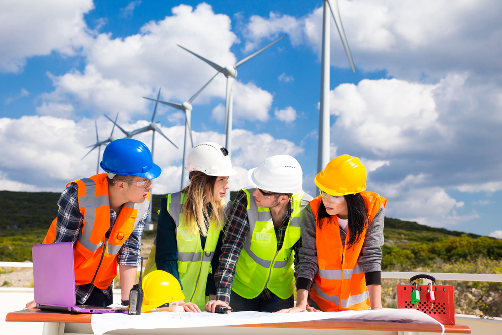 four wind turbine technicians with hardhats look over work plans with wind turbines behind them