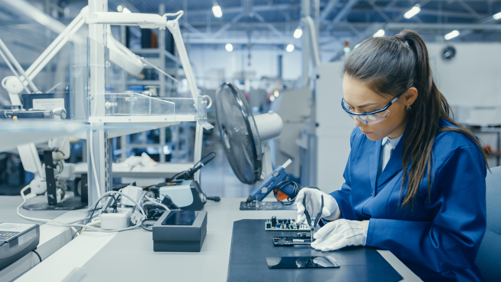 Young female in blue lab coat and safety glasses assembles a circuit board in a high-tech facility, example of electrical technician training