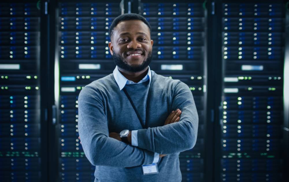 A man poses for a photo with his arms crossed in front of a wall of lit-up servers.