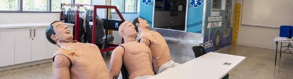 Three CPR mannequins sitting on chairs in a classroom.