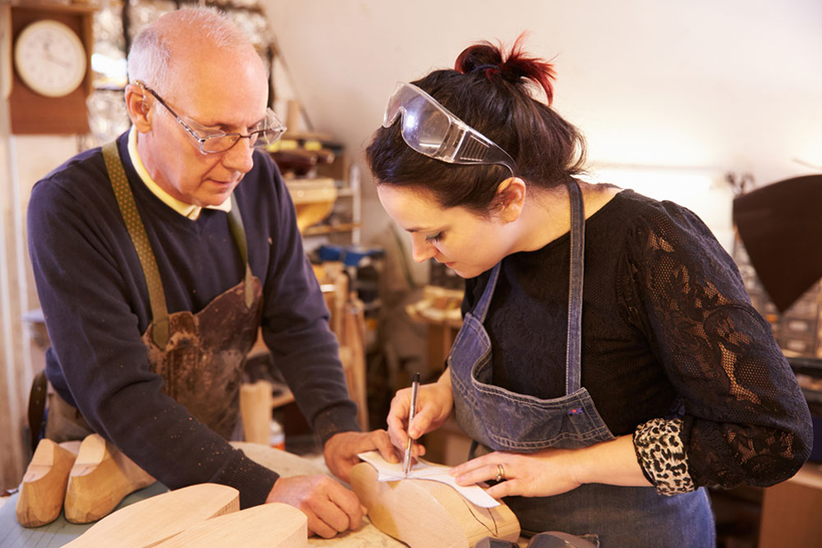 Older male shoemaker teaches young female apprentice in a shop