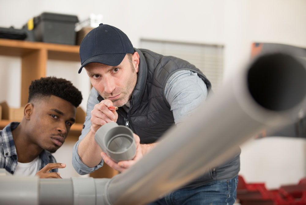 Middle aged white skilled worker trains a young black apprentice on how to align pipes