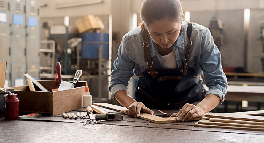 Young female carpenter works with equipment on wooden table in carpentry shop. Example of the benefits of trade school grants and scholarships and how they support young people trying to break into trade jobs.