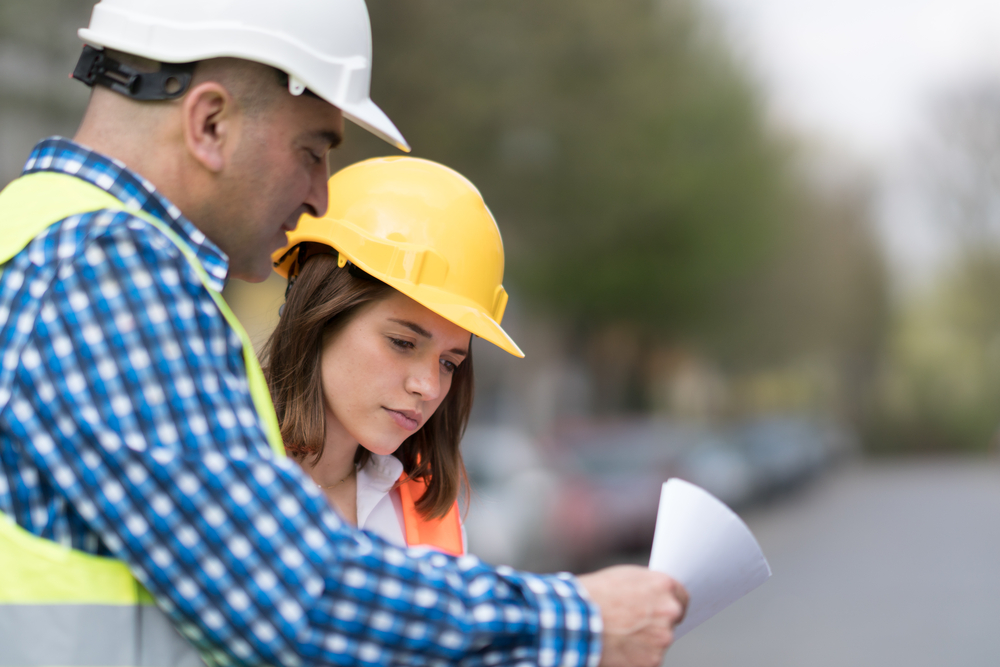 Male and female civil engineers wearing protective vests and hardhats check office blueprints on construction site