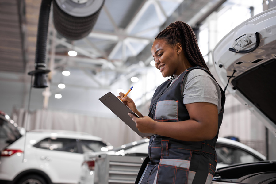 A black, female mechanic writes on a clipboard in a repair shop