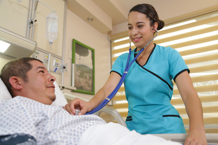 young female Latino nurse takes a male patient's vital signs while he lies in a hospital bed