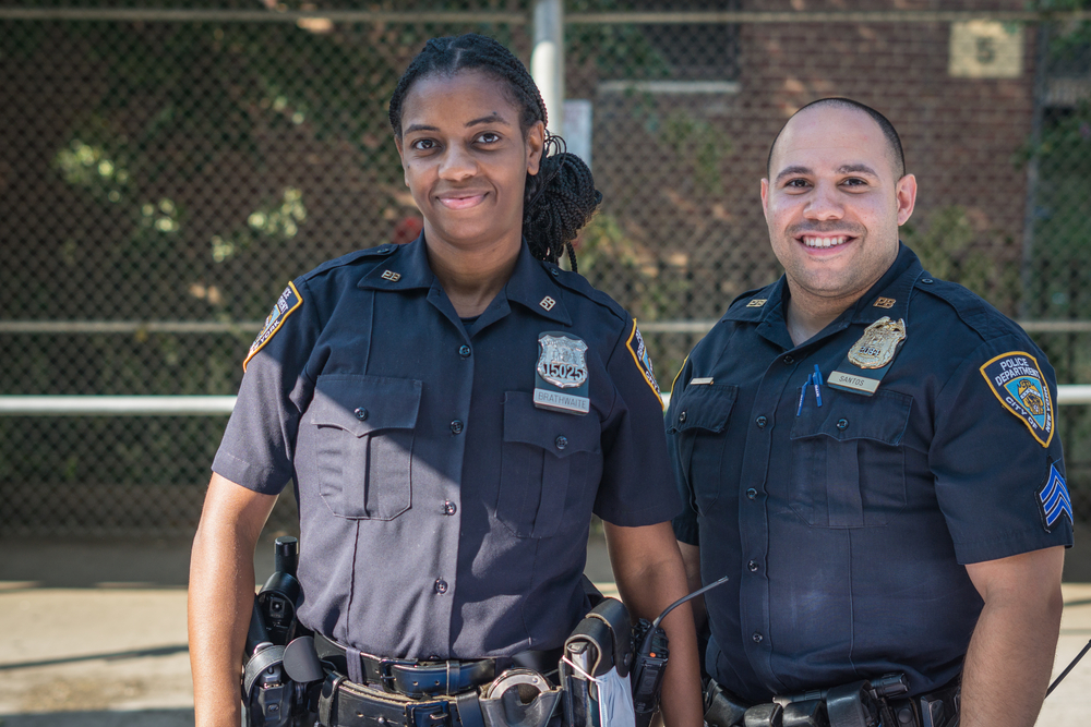 Two NYC police officers in uniform smile at a community event in the Bronx, example of how to become a police officer