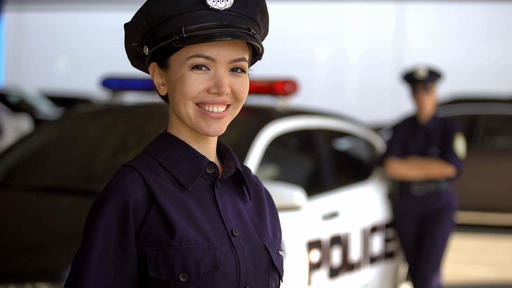 Female police recruit stands in front of a police car with a veteran officer in the background, example of how to become a police officer