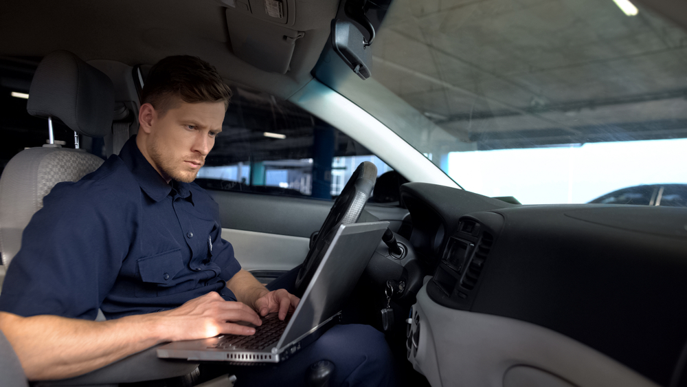 Male police officer works on laptop in car, filling data in crime report, example of how to become a police officer