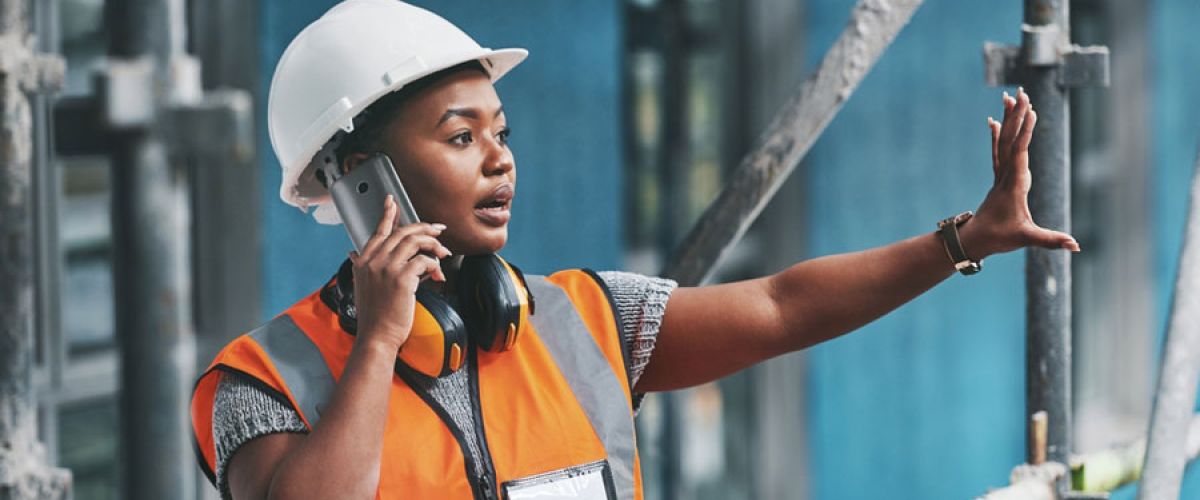 Black female construction worker in an orange safety vest and a hardhat makes a call from a construction site