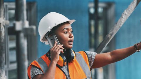 Black female construction worker in an orange safety vest and a hardhat makes a call from a construction site