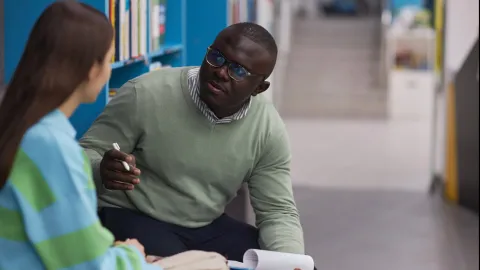 A Black male counselor talks with a female student with long, black hair and a colorful striped sweater