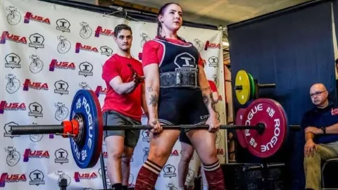 Weightlifter Angeline Deluca stands holding a straight bar with weights on either end