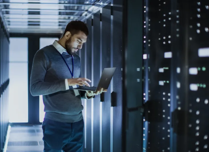 A male network systems administrator runs diagnostics and maintenance testing by a bank of servers