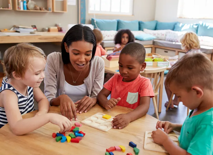 A black female preschool teacher helps three preschool students learn their shapes