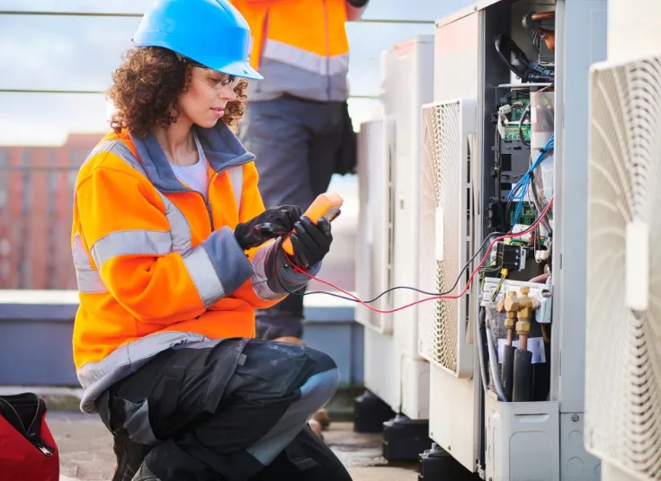 Female HVAC technician in a hardhat and safety gear works on a rooftop HVAC unit