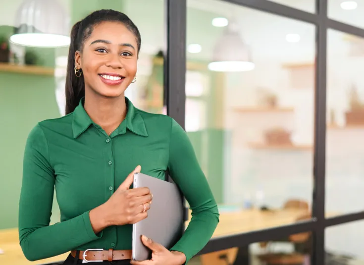 Young Black woman in green shirt smiles at the camera holding a laptop