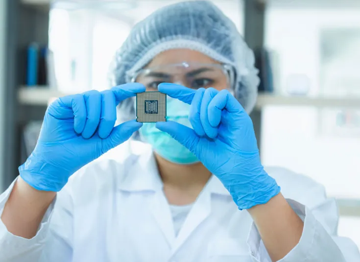 A female semiconductor technician wearing a clean suit examines an integrated circuit in a clean room in a manufacturing facility.