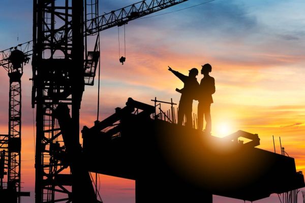 Two silhouetted construction workers stand together on a job site with the sun setting behind them.