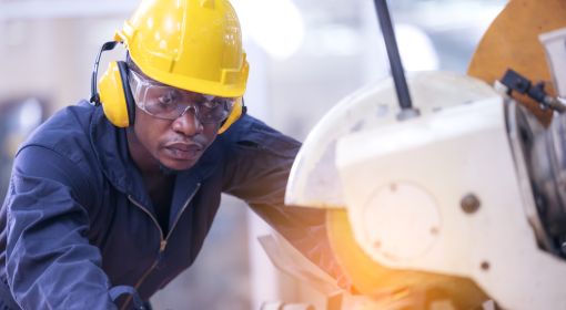 Black male worker wears soundproof headphones and yellow helmet working at a cutting machine