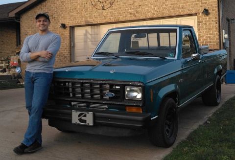 A young man stands in his driveway, leaned up against his turquoise Ford Ranger truck. 