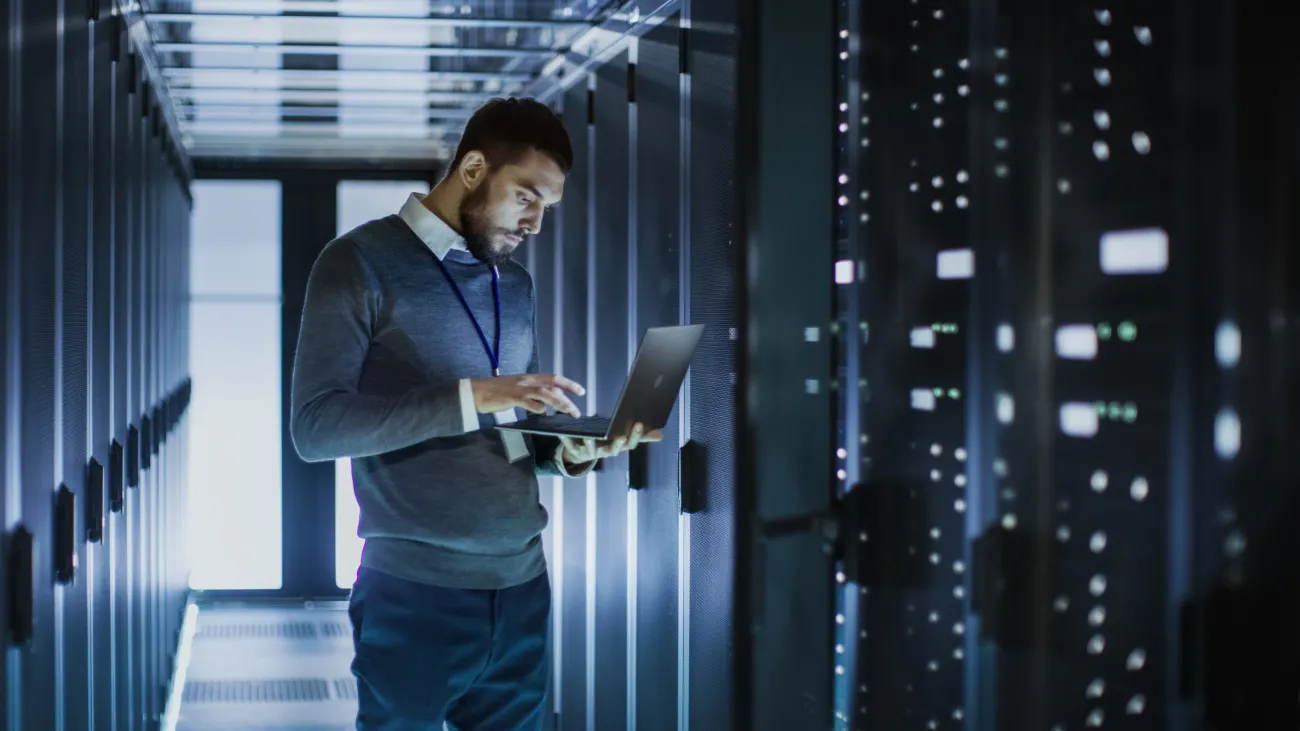 A male network systems administrator runs diagnostics and maintenance testing by a bank of servers