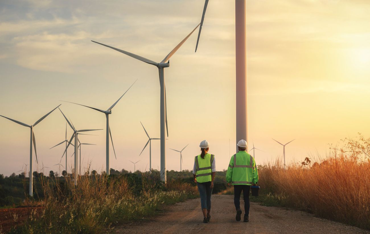 2 wind turbine technicians in safety vests walk toward wind turbines in a field