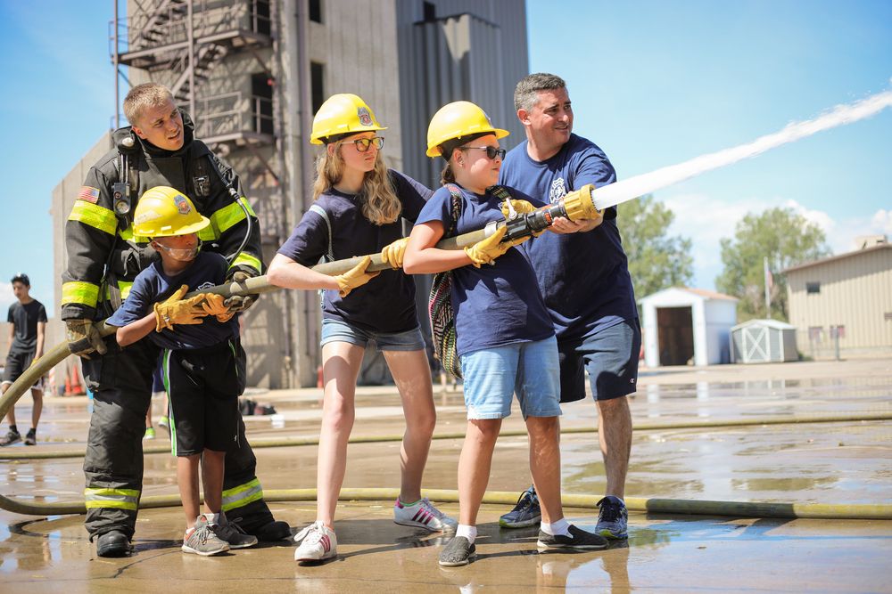Middle school kids with yellow hardhats on shoot water out of a fire hose with help from real firefighters