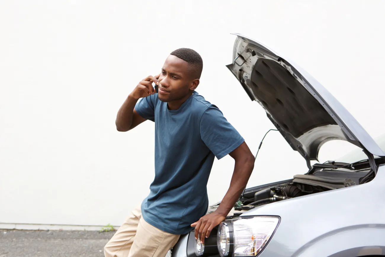 A young man talking on a mobile phone sits on the front of his disabled car with hood open