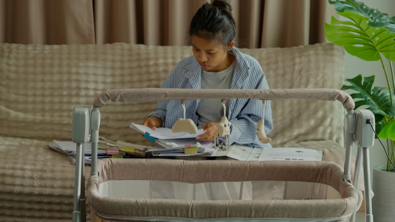 A woman works on homework on the sofa while monitoring a baby's crib