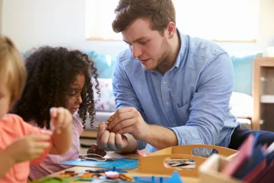Young male preschool teacher works with black girl to solve a puzzle at school. Example of associate degree jobs that are overlooked.