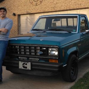 A young man stands in his driveway, leaned up against his turquoise Ford Ranger truck. 