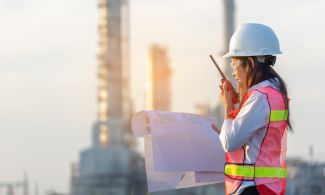 An industrial engineering technician reviews a blueprint while talking on a walkie-talkie in front of a worksite